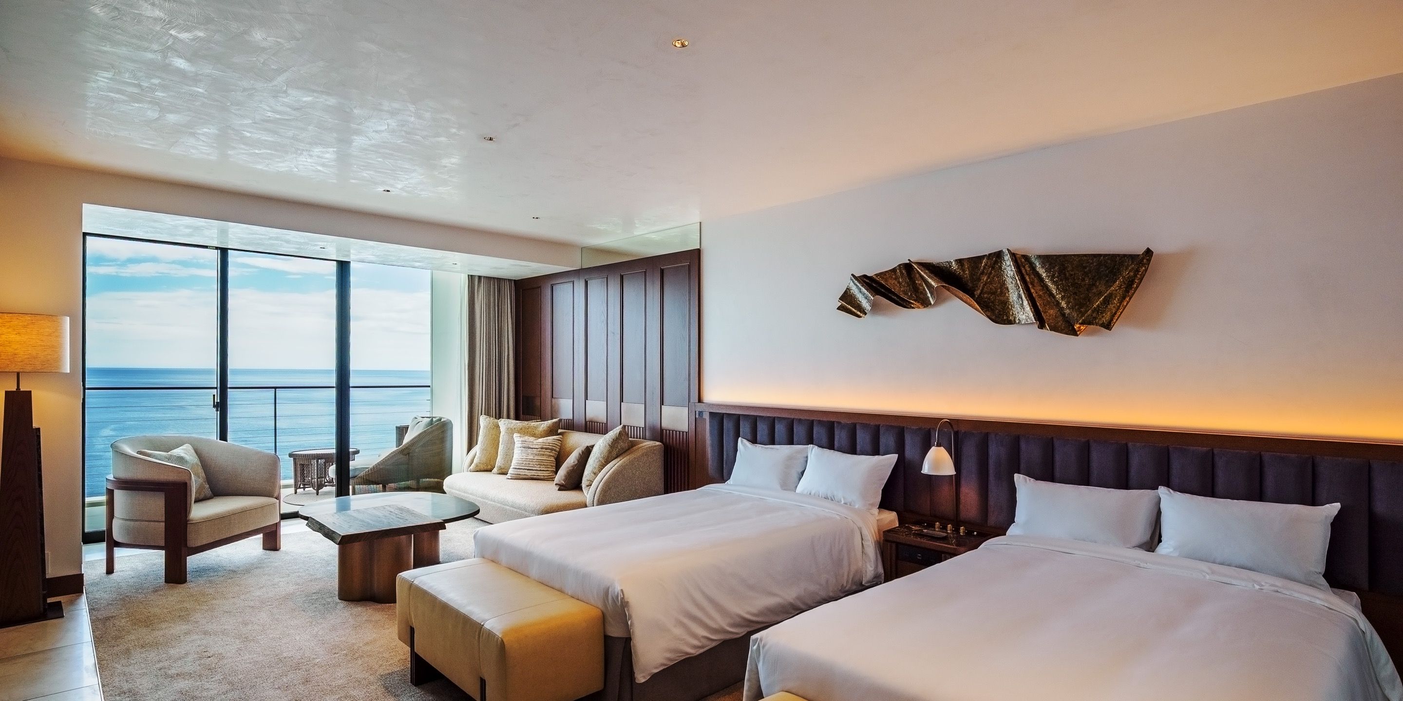 A hotel room with a large window, two beds, a seating area, and a decorative wall piece.