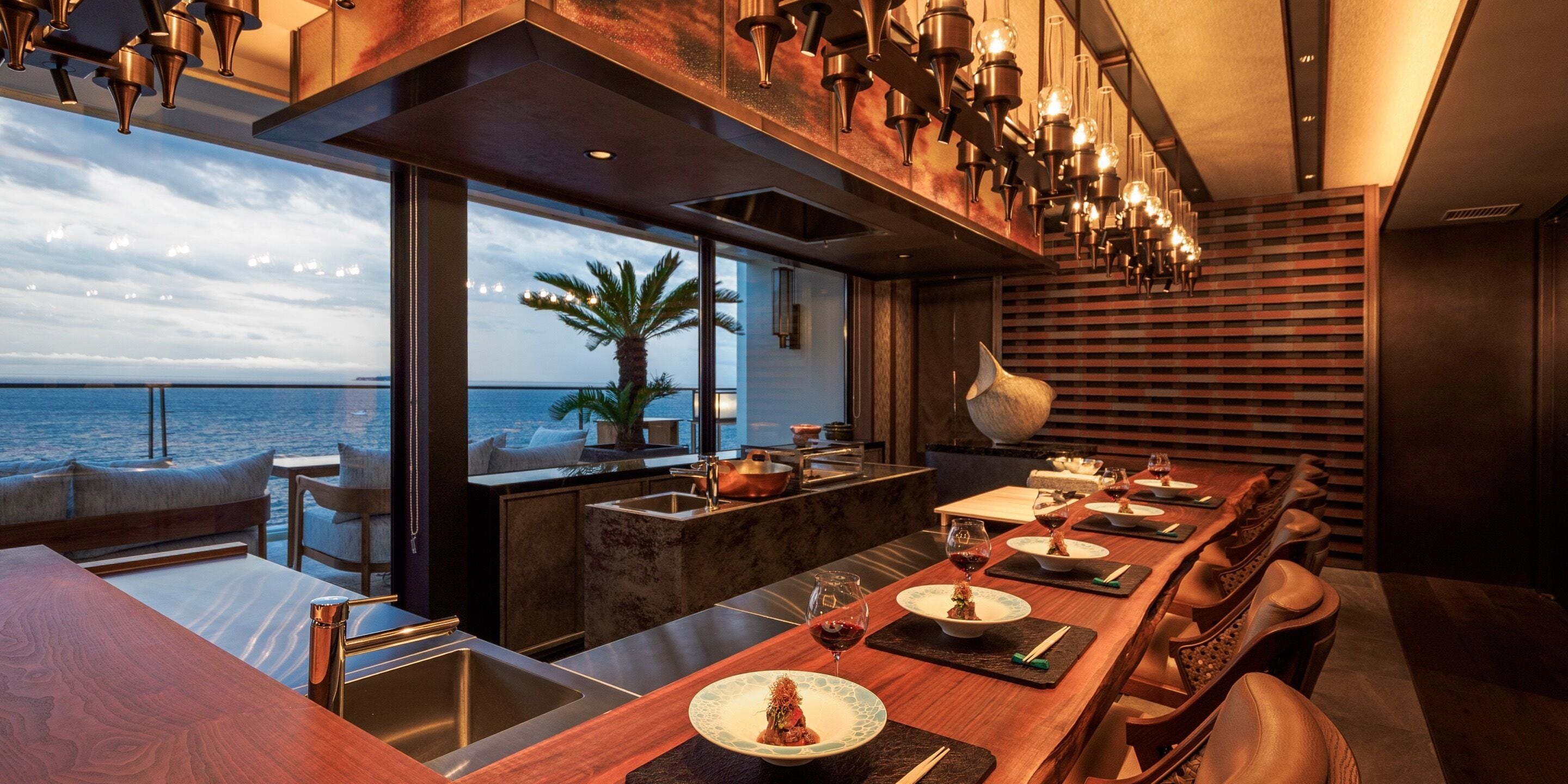 A modern dining area with a view of the sea, a bar with a copper sink, and a long wooden table set for a meal.