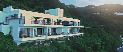 A multi-story modern building with balconies, situated on a hillside with dense greenery.
