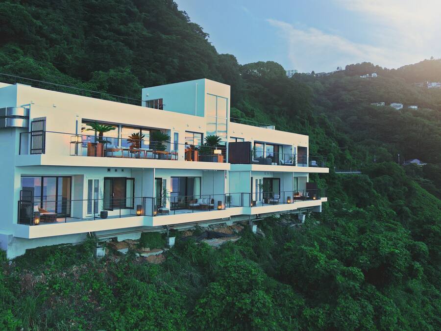 A multi-story modern building with balconies, situated on a hillside with dense greenery.