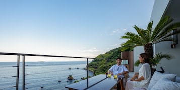 A terrace with a view of the sea, a palm tree, and a dining table with two glasses of a yellow beverage.