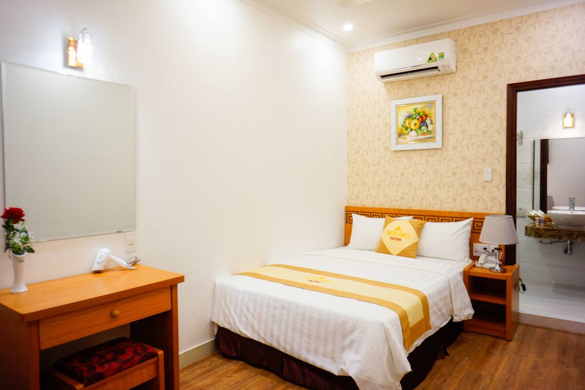 A hotel room with a bed, a bedside table, a mirror, a wall-mounted air conditioner, and a painting.