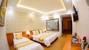 A hotel room with two beds, a wooden floor, a TV mounted on the wall, and a small table with toiletries and a vase of flowers. - Nu Hoang Hotel (Phan Rang)