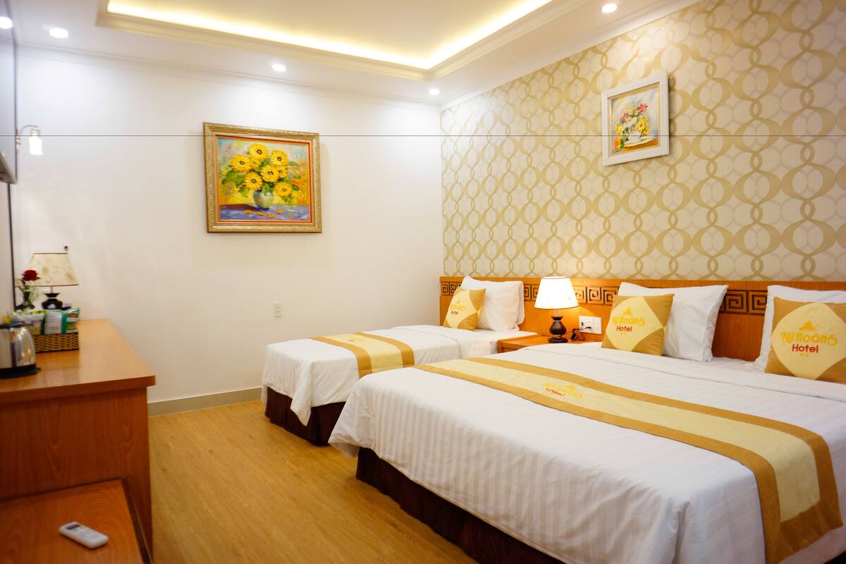 A hotel room with two beds, a desk, and a nightstand with a lamp. There is a painting on the wall and a patterned wallpaper.