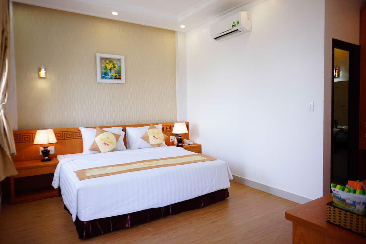 A hotel room with a large bed, two bedside tables with lamps, a wall-mounted air conditioner, and a painting above the bed.