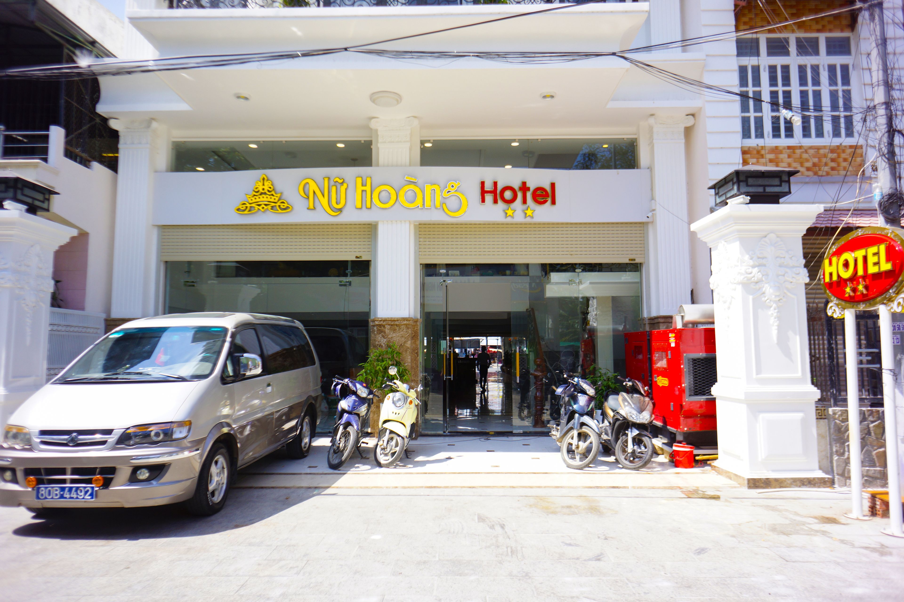A hotel entrance with a sign reading "Nü Hoang's Hotel" and a parked van and scooters outside.