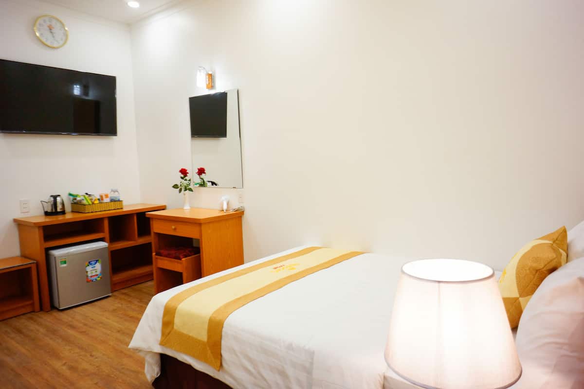 A hotel room with a bed, a desk with a refrigerator, a TV mounted on the wall, and a lamp.