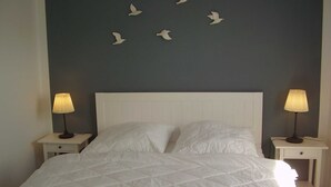 A neatly made bed with a white quilt, two bedside tables with lamps, and a wall with white bird decorations. - Vacation home in Dornumersiel 20-138a - 200-138a (Dornum)