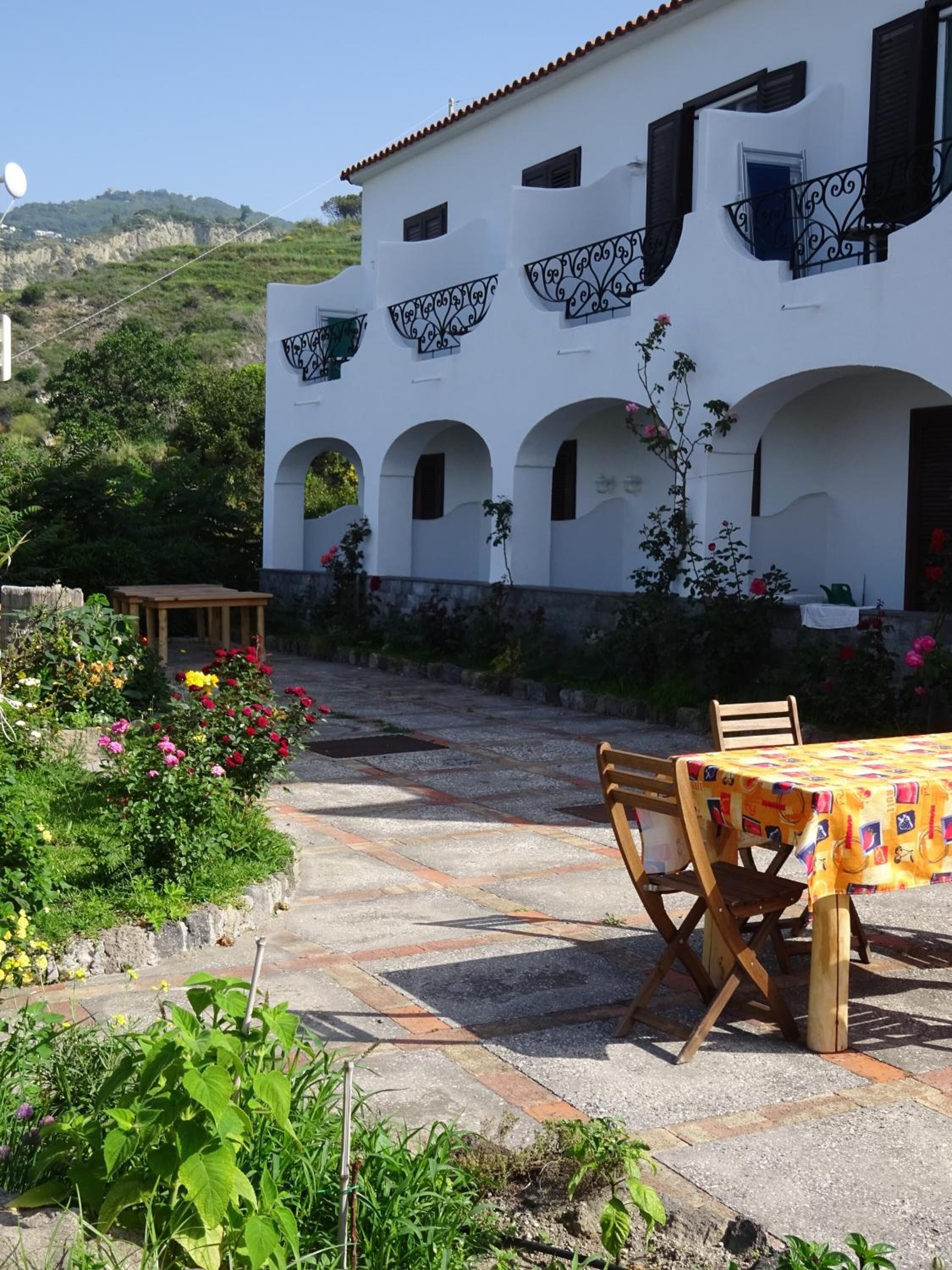 A white building with arches and balconies, a patio with a table and chairs, and a garden with flowers.