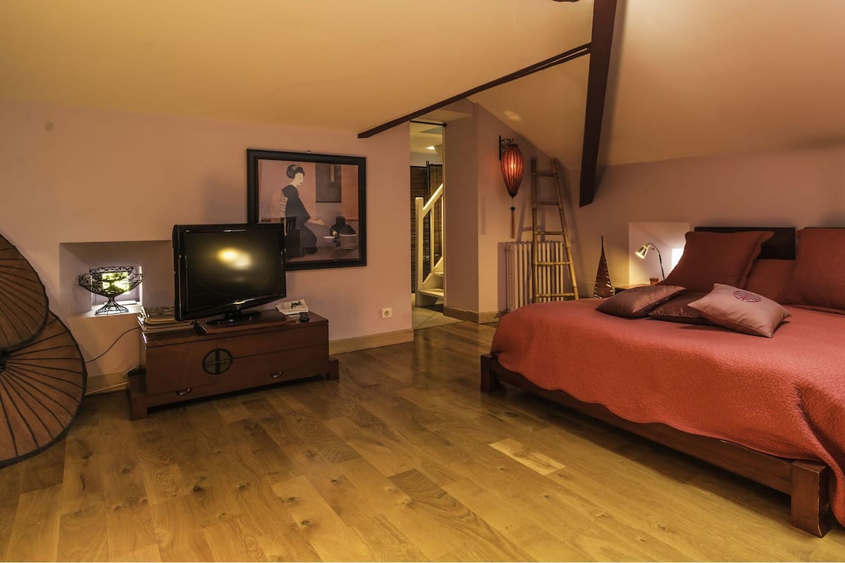 Een slaapkamer met een houten vloer, een bed met rode beddengoed, een televisie op een houten dressoir en een lijst met een foto aan de muur.