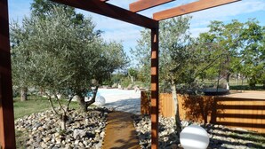 Una terraza de madera con un sendero de piedra que conduce a una zona de piscina, rodeada de olivos y una valla de madera.