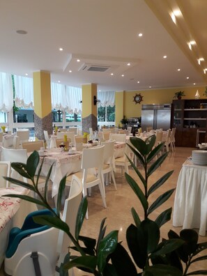 A dining area with tables set for a meal, chairs, a refrigerator, and a clock on the wall. - Hotel Maxim (Misano Adriatico)