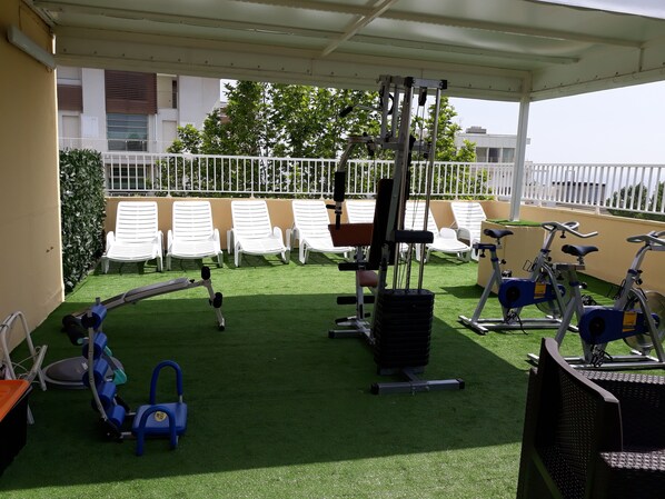 Zona de gimnasio al aire libre con equipo de ejercicios, incluyendo una máquina de remo, una prensa de banca y bicicletas estáticas, rodeada de sillones blancos.