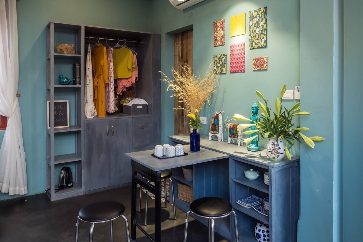 A compact kitchen with a grey cabinet, a small dining table with stools, and a wall-mounted shelf with various items.