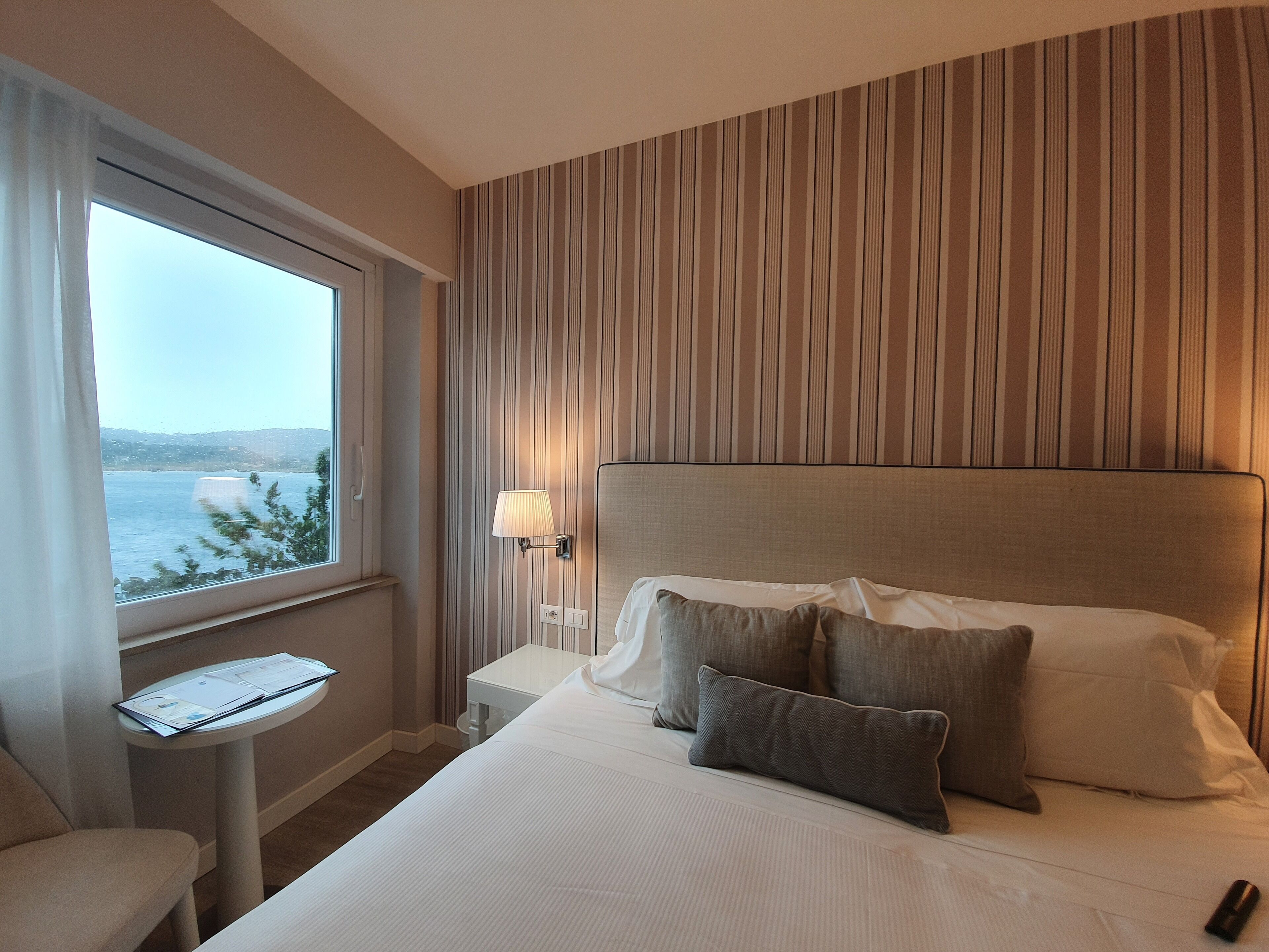 Habitación de hotel con cama, mesita de noche con lámpara, una silla y ventana con vista al mar.