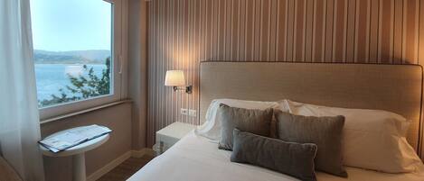 A hotel room with a bed, a bedside table with a lamp, a chair, and a window with a view of the sea.