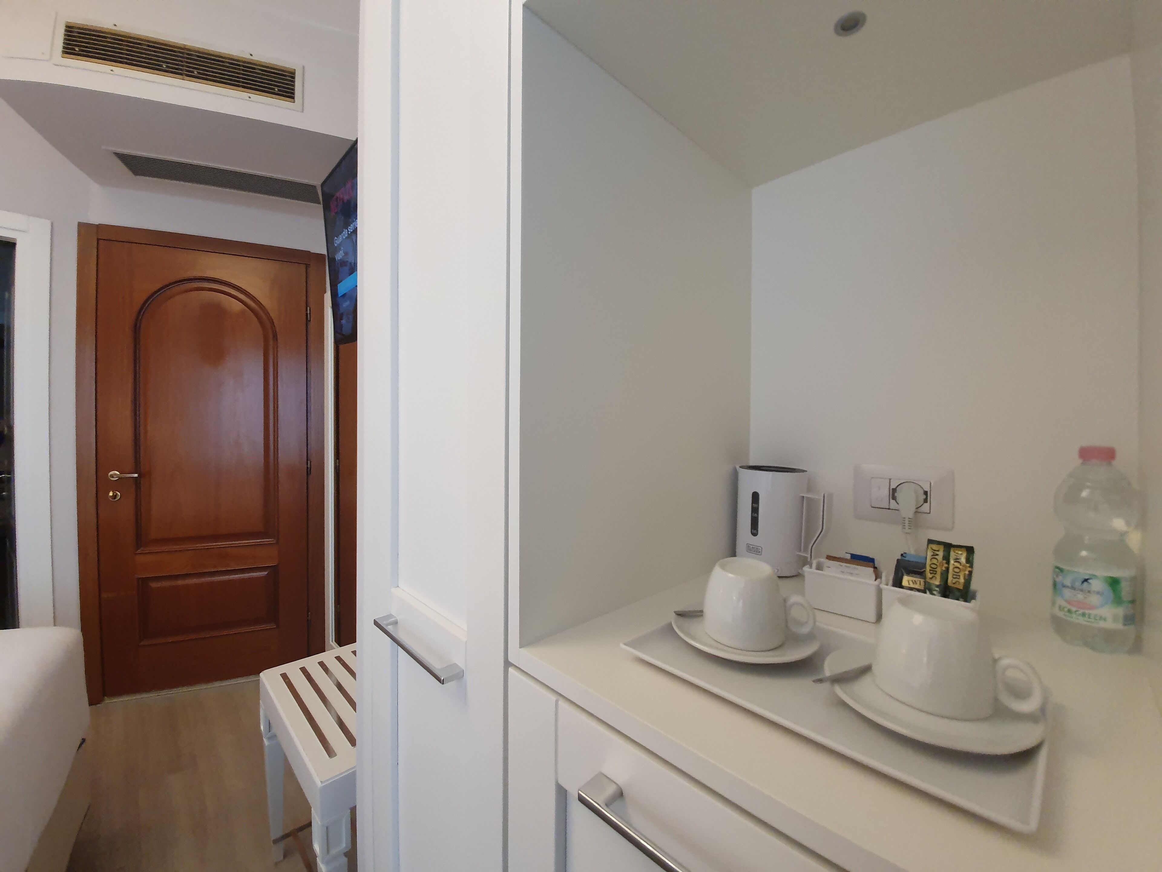 A hotel room with a wooden door, a white sink with a mirror, a white countertop with a kettle, a water bottle, and a small table with a chair.