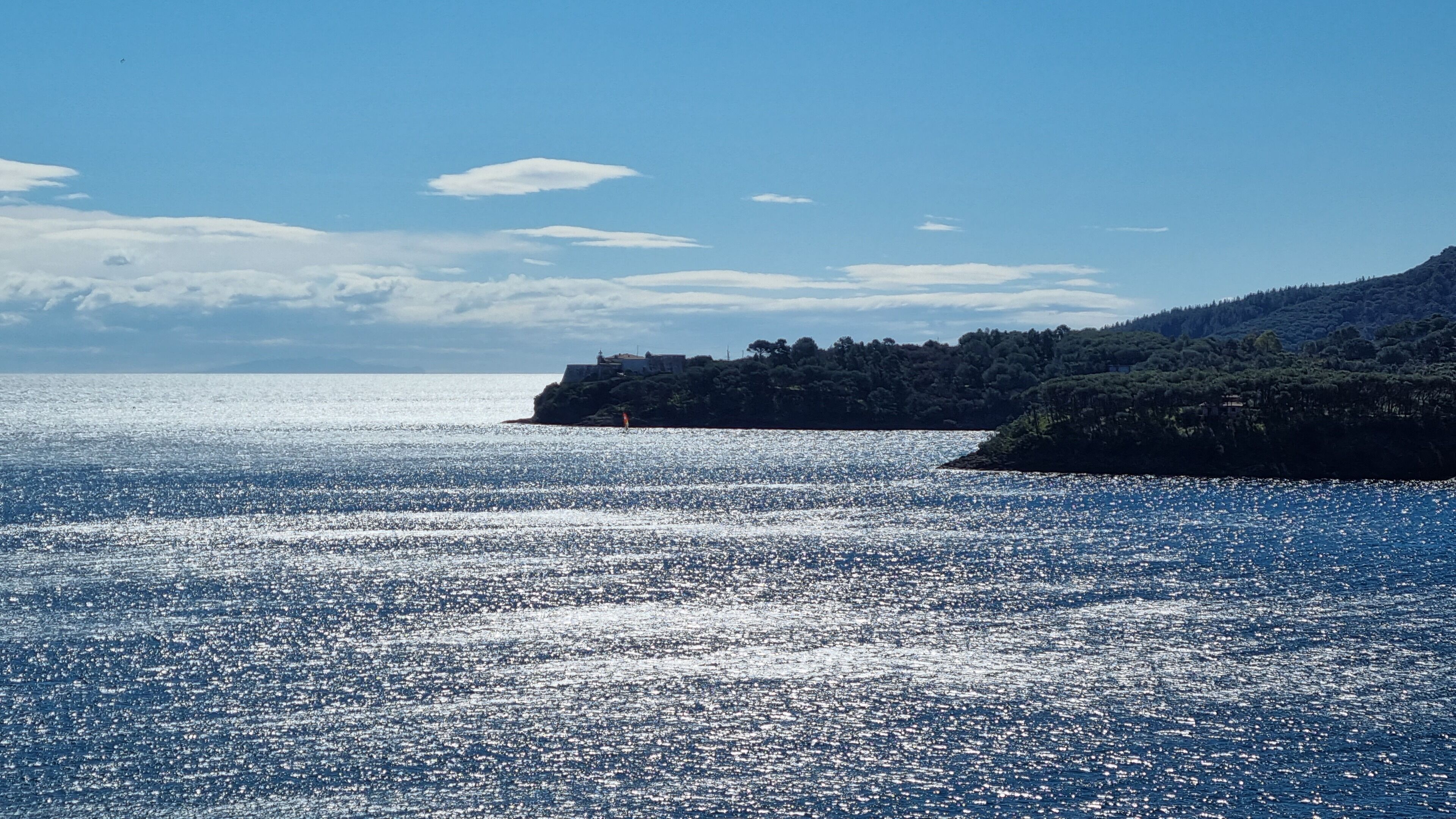 A coastal landscape with a clear sky, calm sea, and a forested hill.