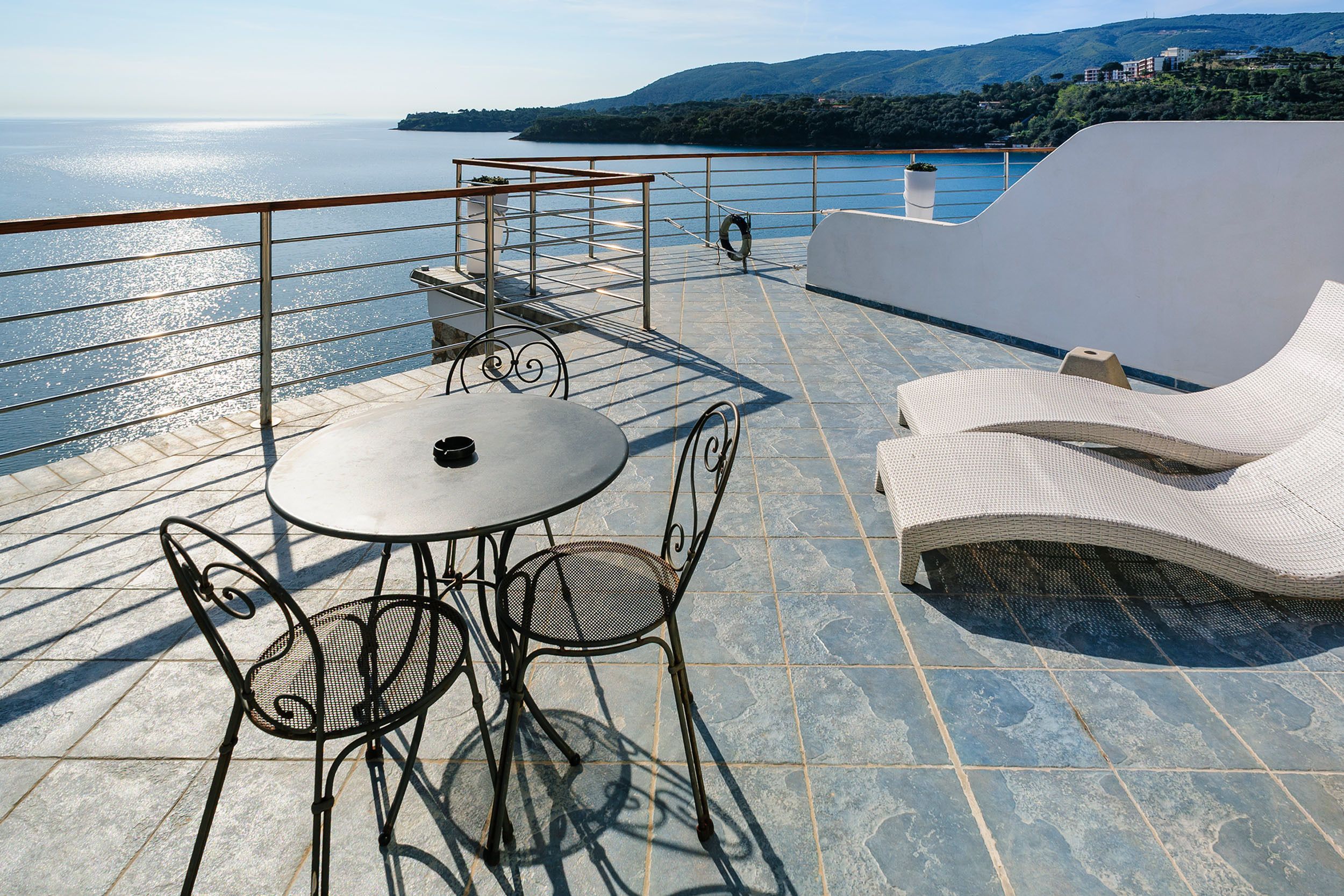 Eine Terrasse mit Tisch und Stühlen, ein Liegestuhl und ein Geländer mit Blick auf ein Gewässer und Hügel.