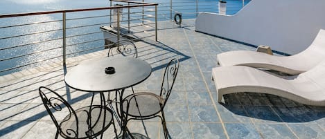 Eine Terrasse mit Tisch und Stühlen, ein Liegestuhl und ein Geländer mit Blick auf ein Gewässer und Hügel.