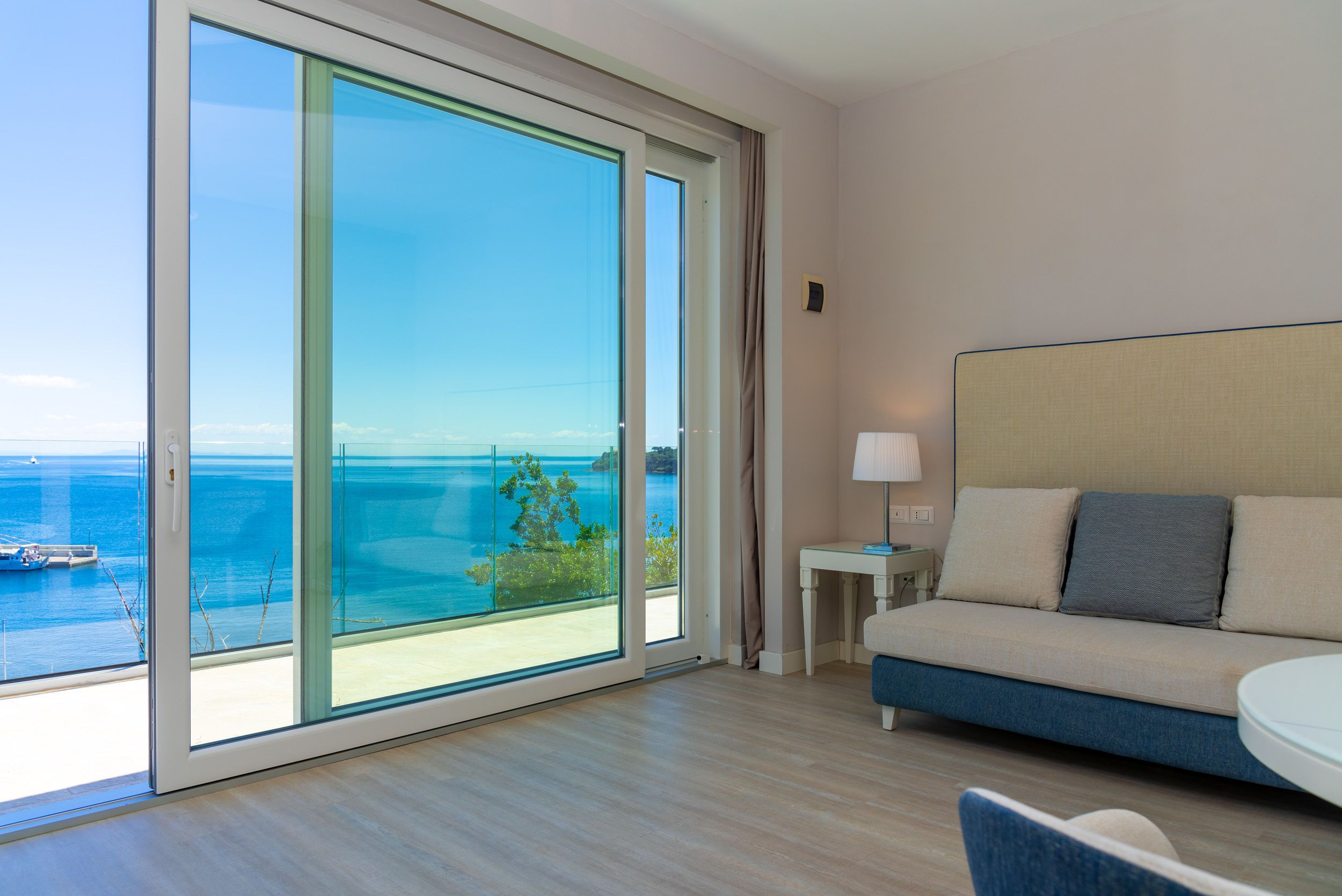 Ein Hotelzimmer mit einer großen Schiebeglasdoor zum Balkon mit Blick auf das Meer und Boote. Es gibt ein Bett mit Kopfteil, einen kleinen Tisch mit einer Lampe und einen Sessel.