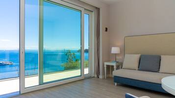 Ein Hotelzimmer mit einer großen Schiebeglasdoor zum Balkon mit Blick auf das Meer und Boote. Es gibt ein Bett mit Kopfteil, einen kleinen Tisch mit einer Lampe und einen Sessel.