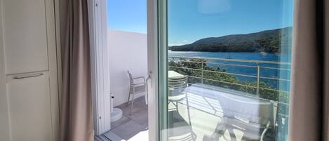 A balcony with a clear view of a bay, a sailboat, and a hilly landscape.