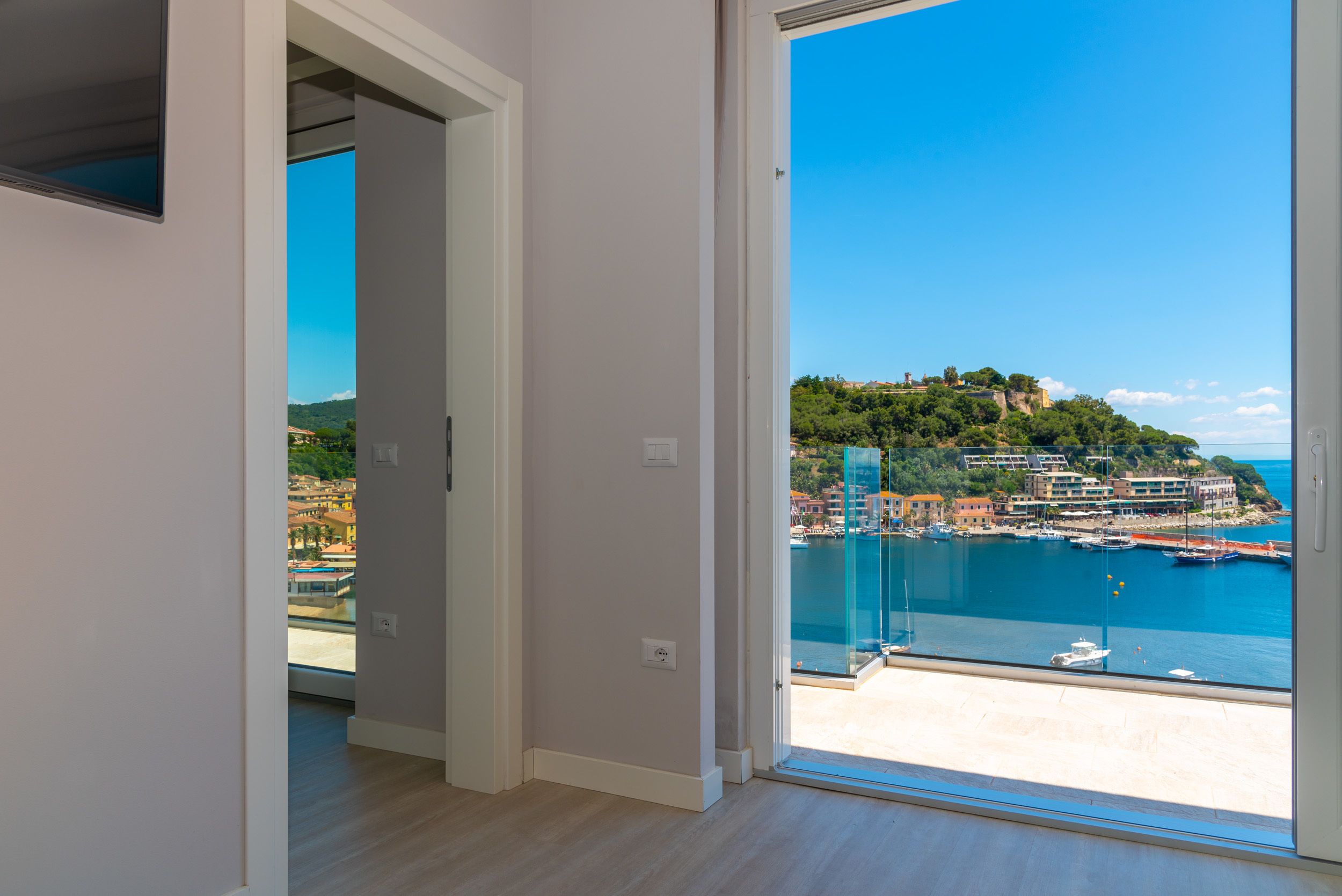 A room with a large sliding glass door leading to a balcony with a view of a coastal town and marina.