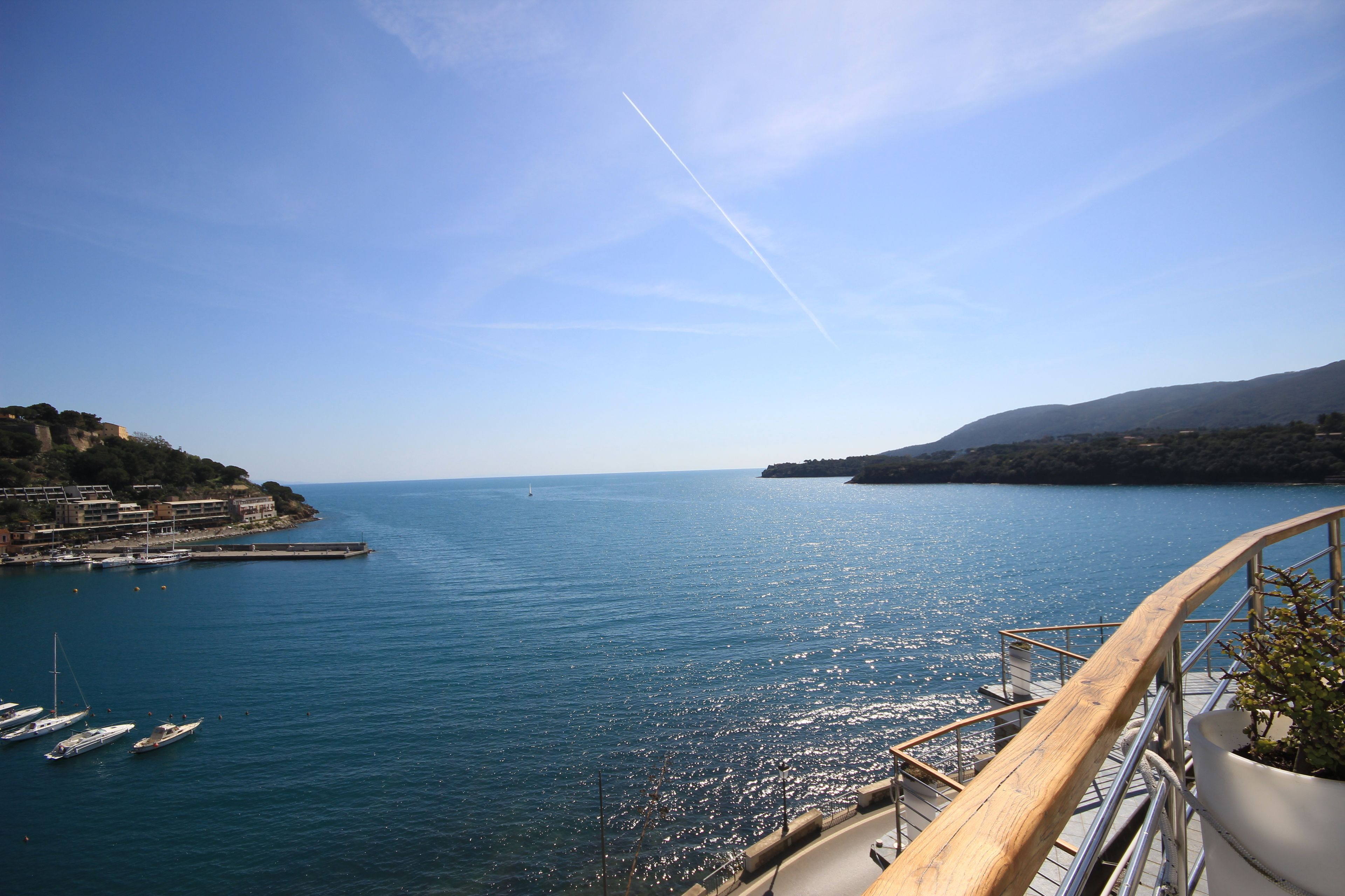 A coastal view with boats docked at a pier, a clear sky, and a mountainous backdrop.