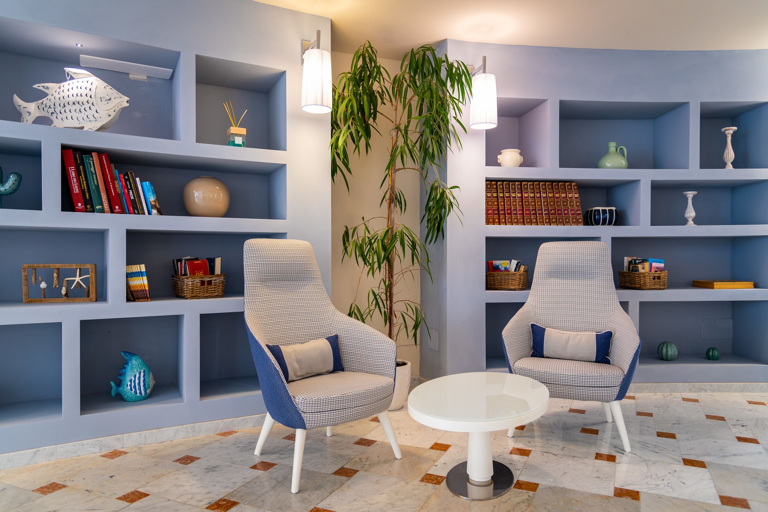 A modern living room with two armchairs, a round table, and a large plant. Shelves with books, decorative items, and a fish sculpture are visible.