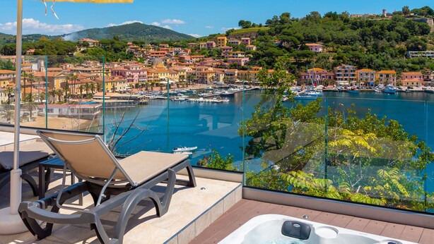 A rooftop terrace with a hot tub, lounge chairs, and an umbrella, overlooking a marina and coastal town. - Hotel Plaza (Porto Azzurro)