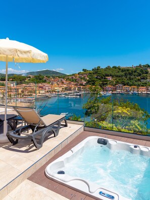A rooftop terrace with a hot tub, lounge chairs, and an umbrella, overlooking a marina and coastal town. - Hotel Plaza (Porto Azzurro)