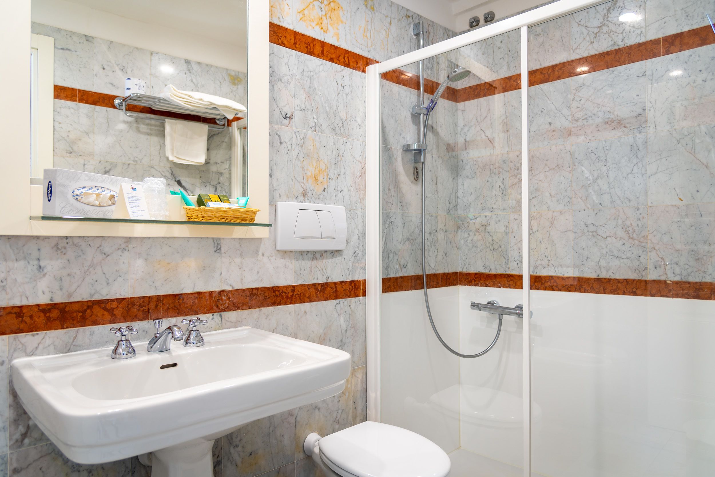 A bathroom with a white sink, a toilet, a glass-enclosed shower, and a marble-tiled wall with orange accents.