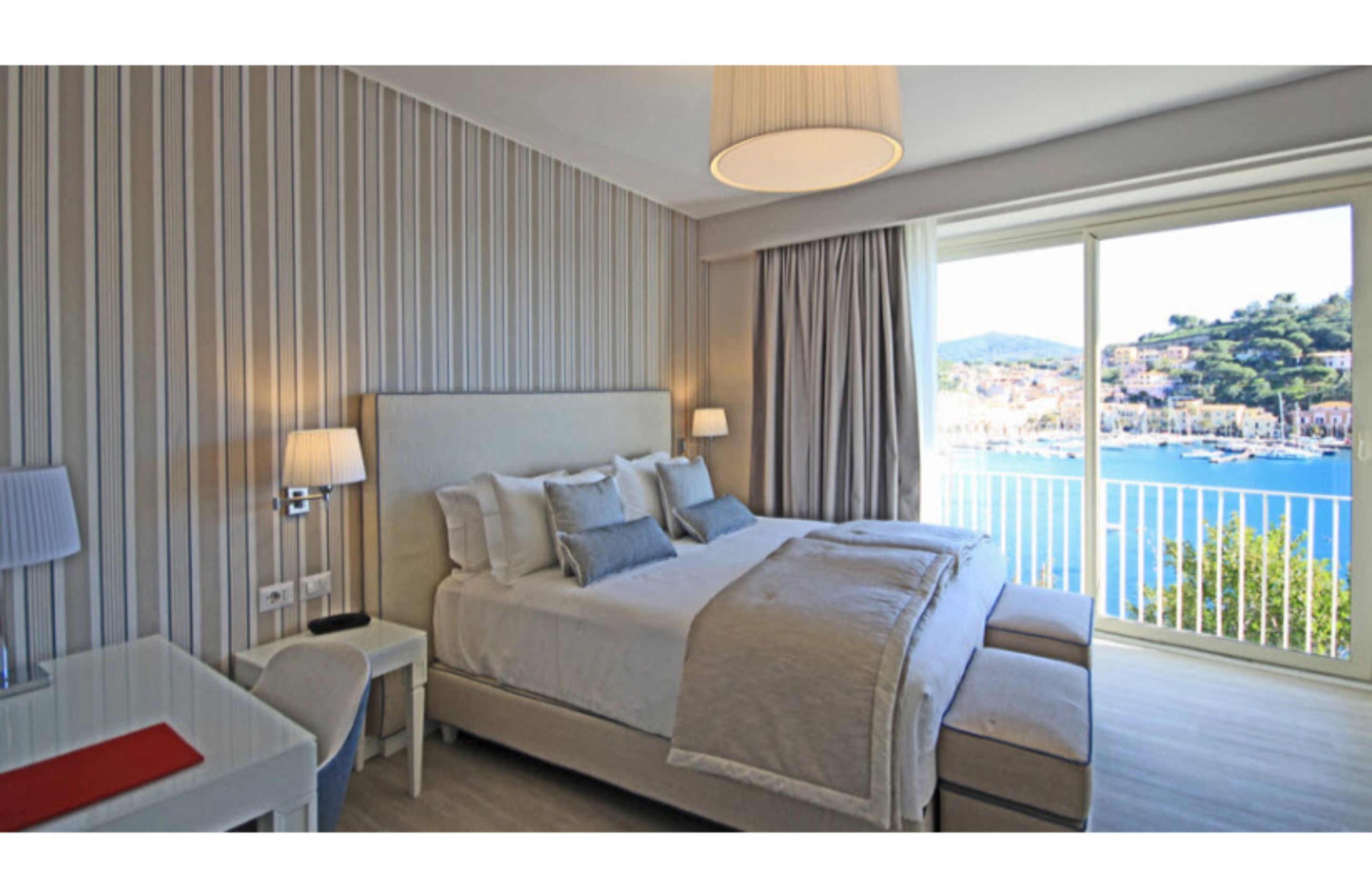 A hotel room with a large bed, two bedside tables, a desk, and a view of a coastal town.