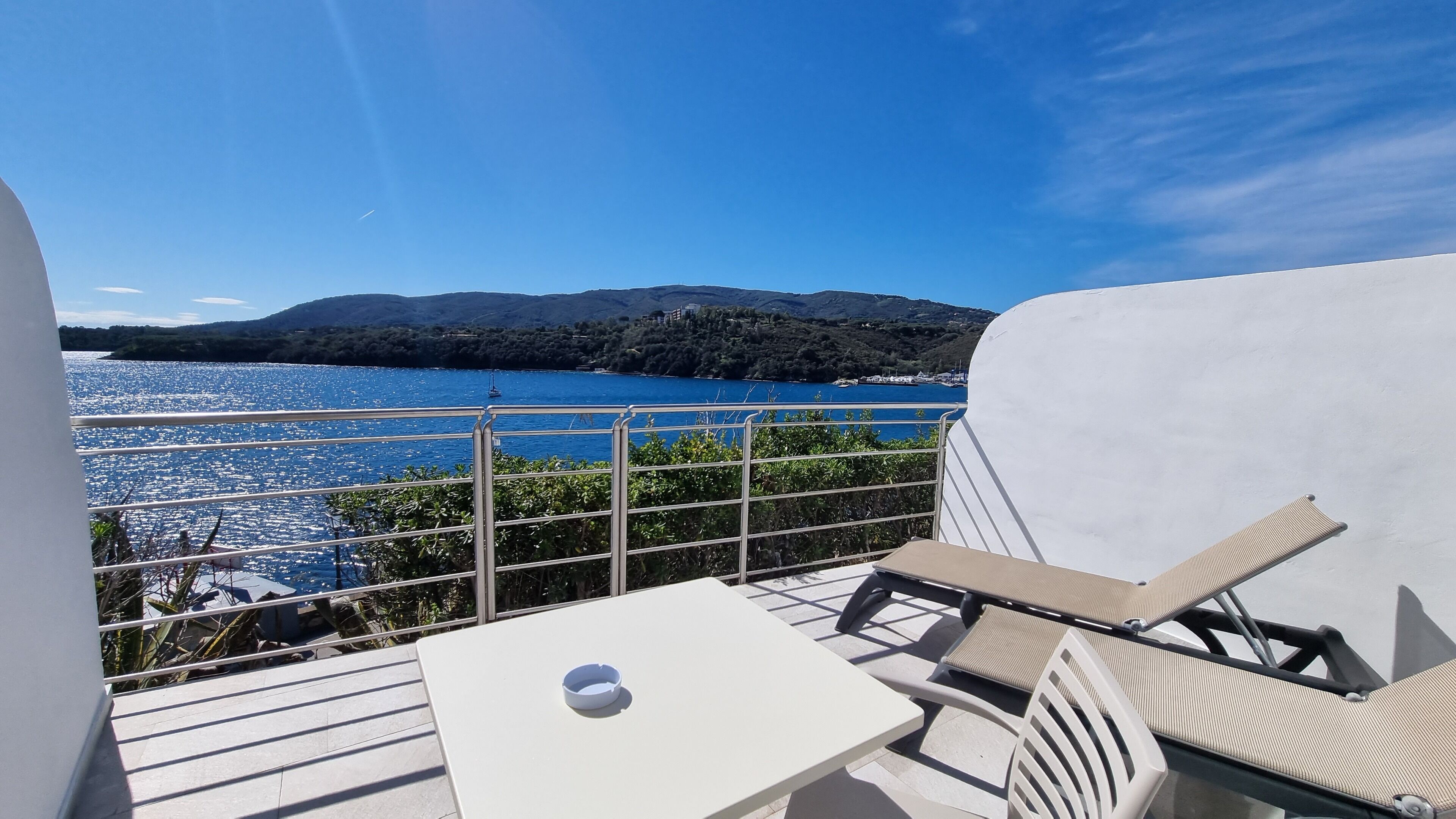 Una terraza con una mesa blanca y dos sillas, con vista a un cuerpo de agua y colinas.