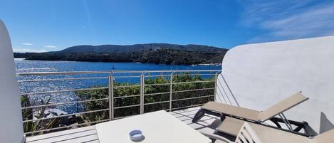 Una terraza con una mesa blanca y dos sillas, con vista a un cuerpo de agua y colinas.