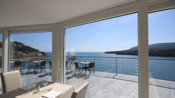 A dining area with a view of the sea and hills, featuring tables and chairs, a potted plant, and a menu on the table.