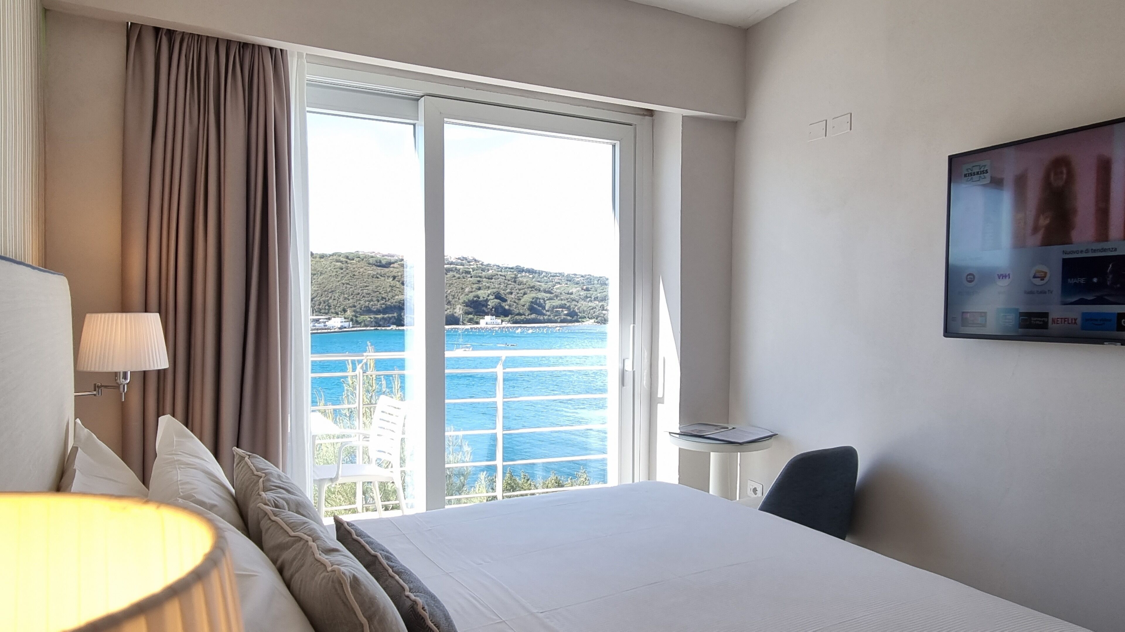 Une chambre d’hôtel équipée d’un lit, d’un bureau, d’une chaise, d’une télévision et offrant une vue sur la mer et les collines.