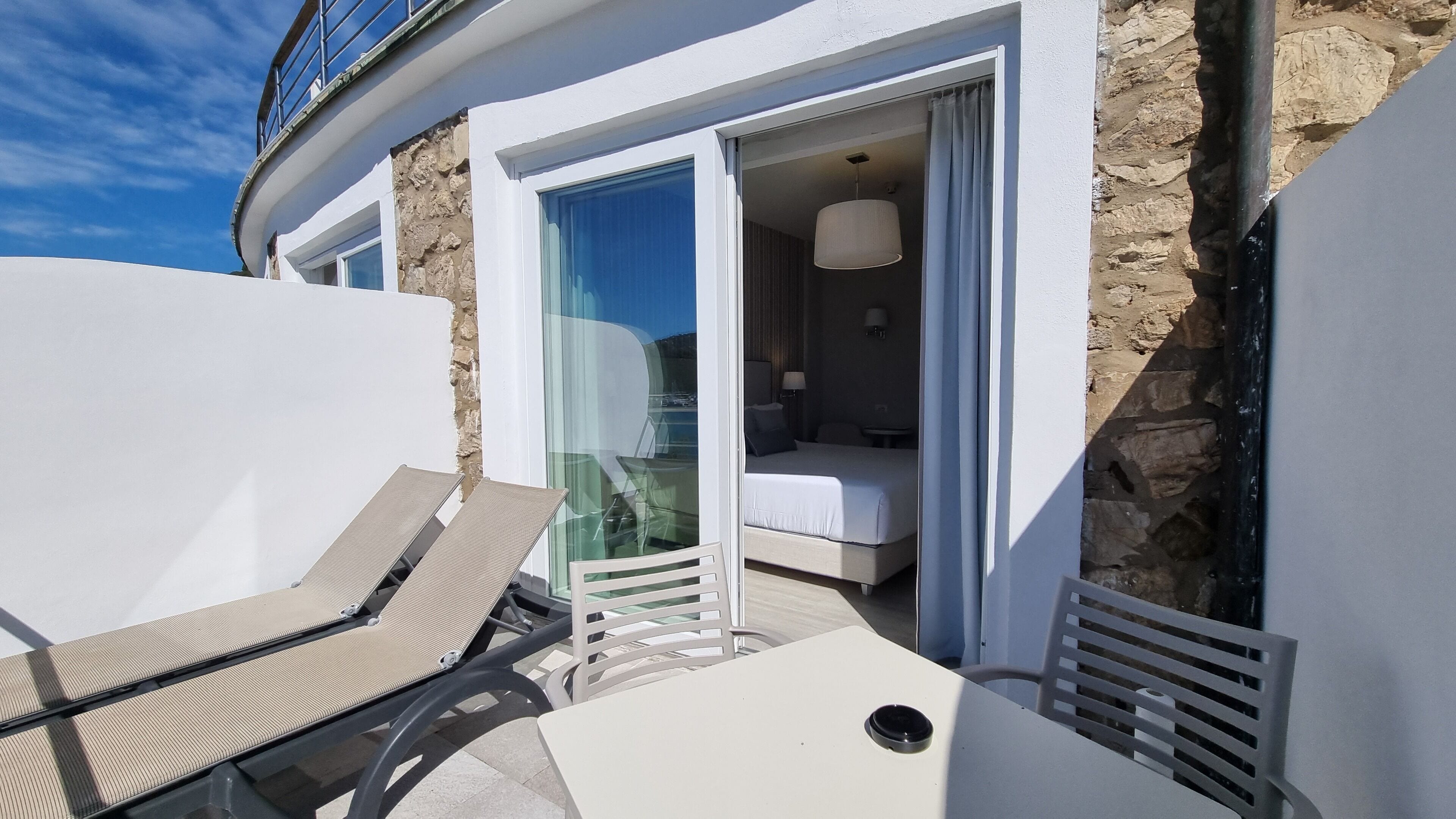 Un balcon avec un fauteuil et une table, une chambre à coucher avec un lit et une table de chevet, et un mur en pierre.