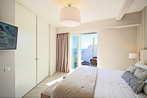 A modern hotel room with a large bed, a desk, and a view of the sea through the balcony doors.