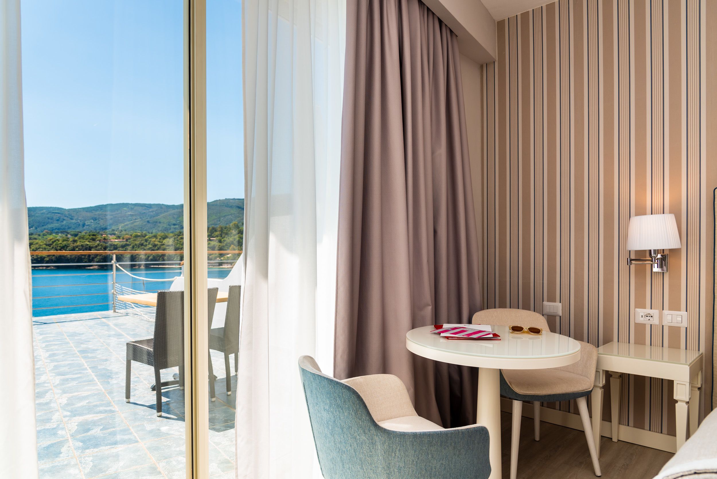 A hotel room with a balcony, a round table with two chairs, a bed, and a view of the sea and hills.