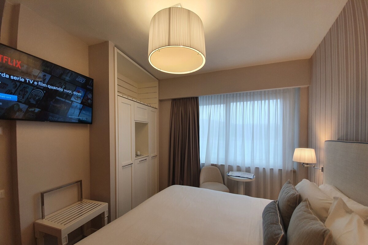 Una camera d'albergo con un letto, un comodino, una lampada, una TV e vista sull'esterno dalla finestra.