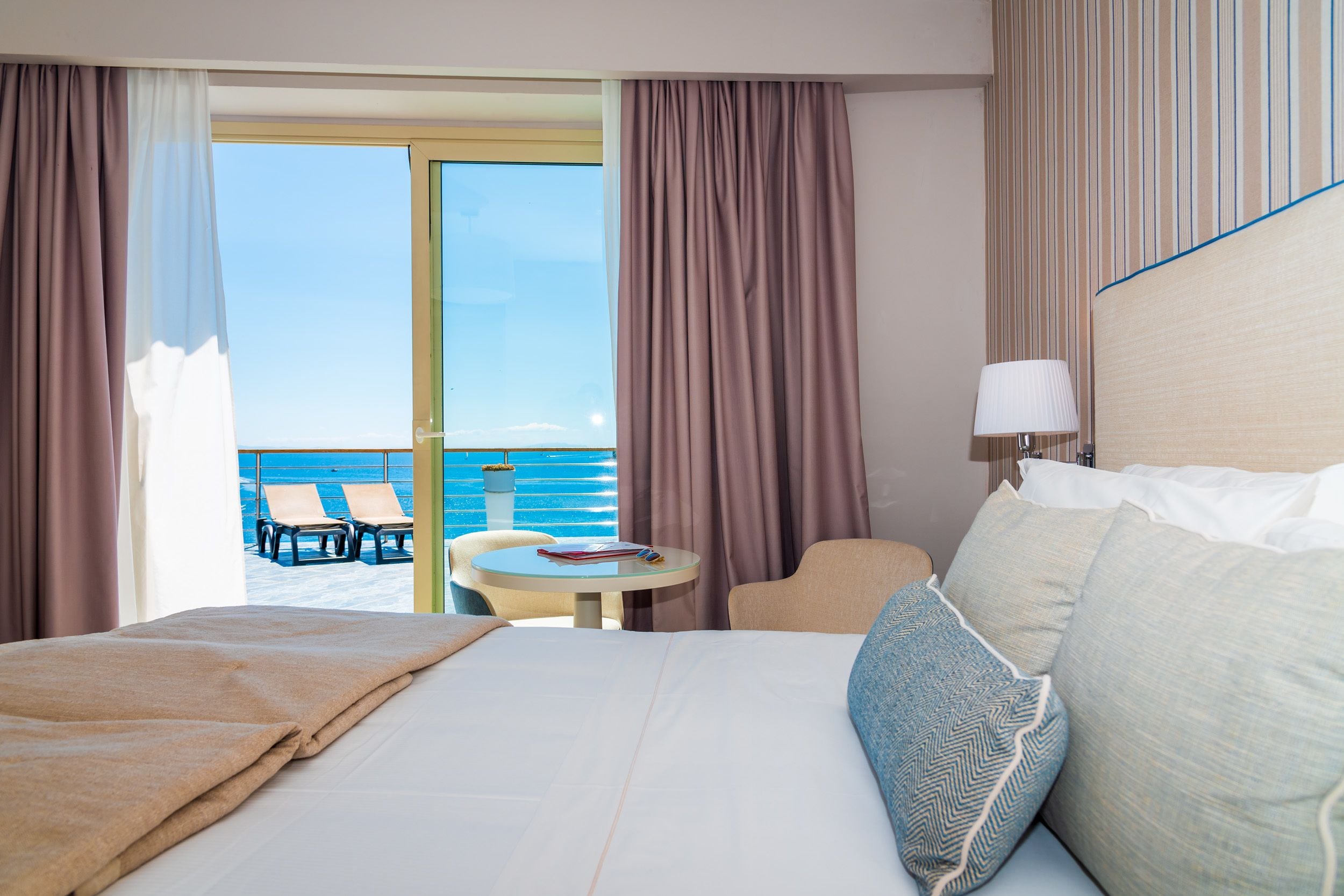 A hotel room with a bed, a small table, a chair, and a view of the sea.