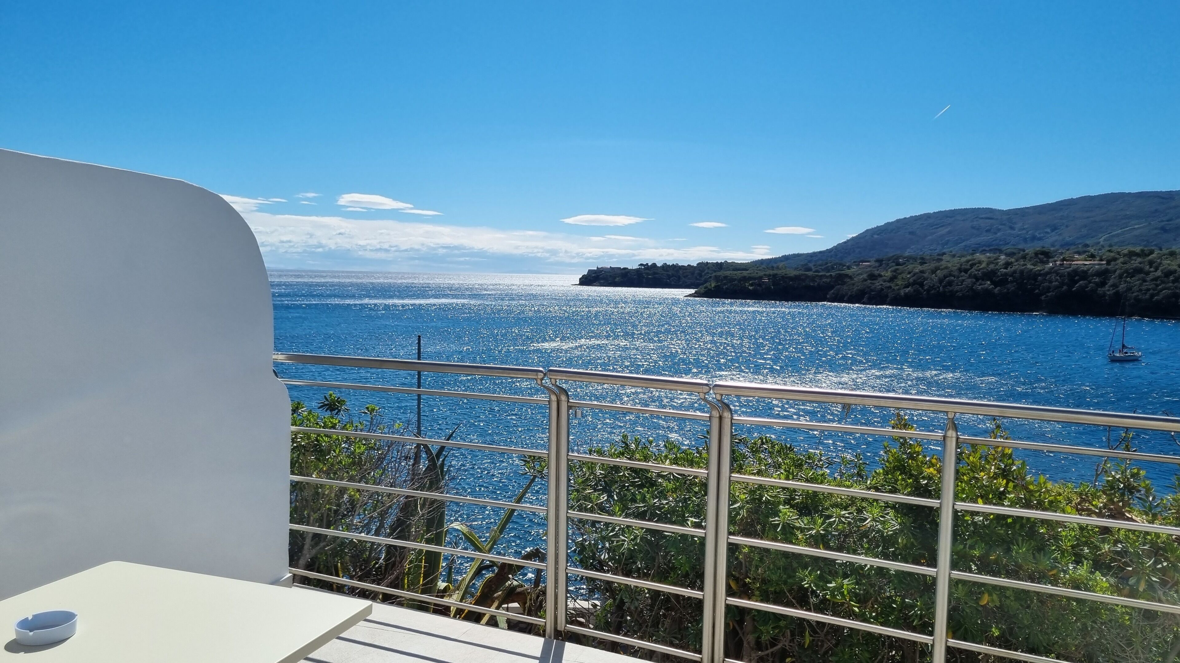 Un balcón con vista a un mar tranquilo, un velero y un paisaje montañoso.