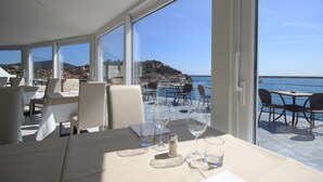 A dining area with tables and chairs, large windows with a view of the sea and a distant hill. - Hotel Plaza (Porto Azzurro)