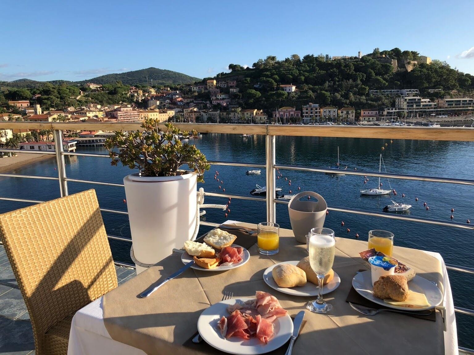 Une table dressée avec de la nourriture et des boissons, donnant sur une marina où sont amarrés des bateaux et sur une ville côtière.