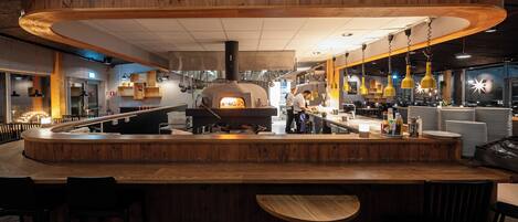 A restaurant with a central wood-burning oven, a curved wooden bar, and hanging yellow pendant lights.