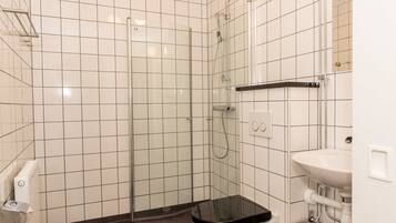Ett badrum med vita kakelväggar, en duschkabin i glas, en svart toalettstol och ett vitt handfat.