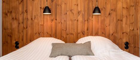 Una cama con almohada a rayas y una colcha, frente a un cabecero de madera con dos lámparas colgantes.