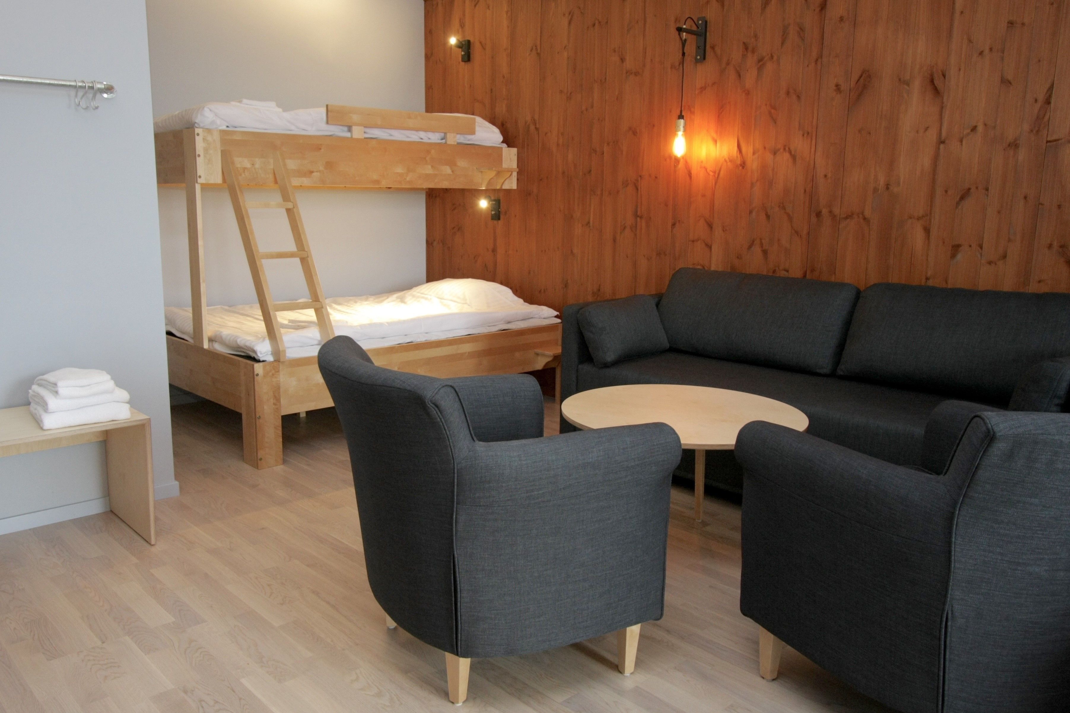 Una habitación con una cama litera, un sofá, dos sillones, una mesita redonda y una pared de madera.